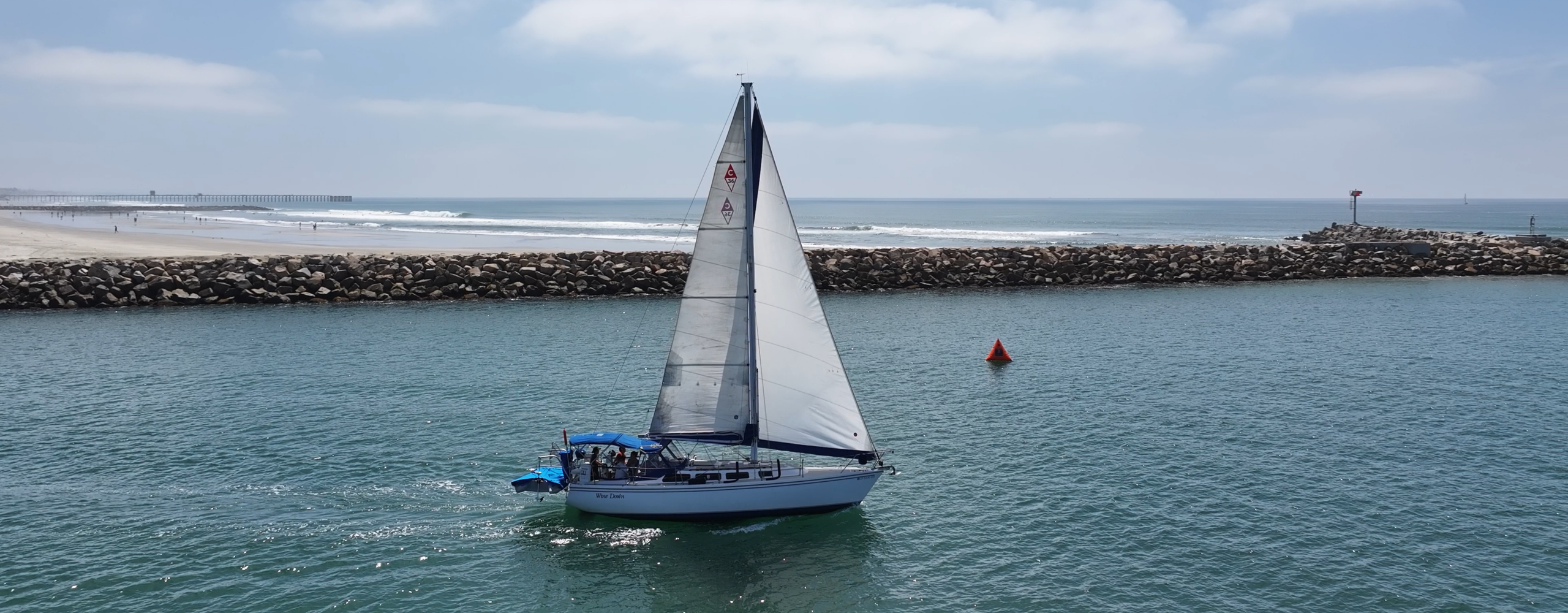 Cruise Oceanside waters on a classic Catalina sailboat. A sailboat passes in front of a jetty on a sunny day